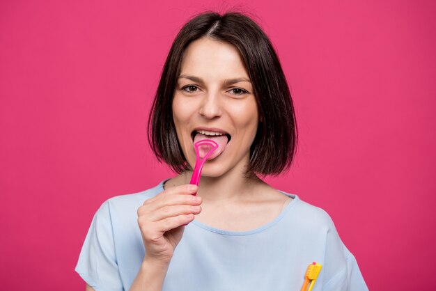 Hermosa mujer joven feliz con raspador de lengua sobre fondo rosa en blanco