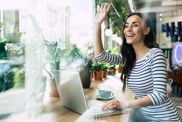 Hermosa mujer joven feliz usando una computadora portátil y bebiendo una taza de café en la cafetería mientras almuerza