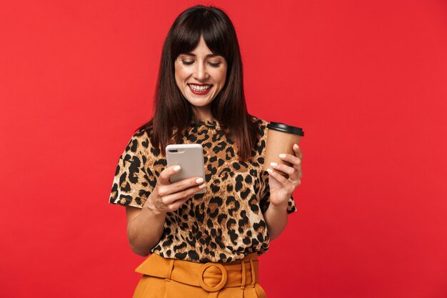 Hermosa mujer joven feliz vestida con camiseta estampada de animales posando aislada sobre pared roja tomando café con teléfono móvil.