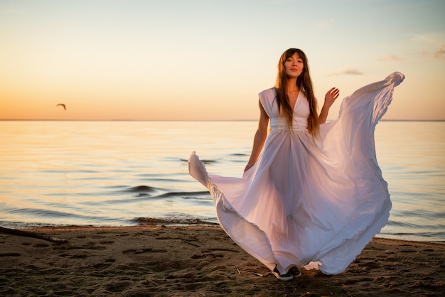 Hermosa mujer joven feliz con un vestido largo blanco en la orilla del mar al atardecer se encuentra