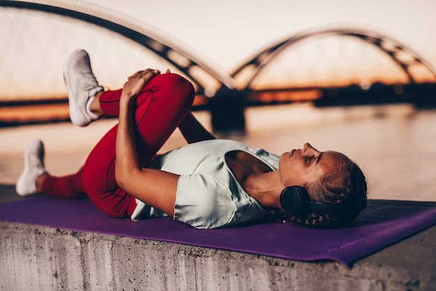 Hermosa mujer joven y en forma en buena forma ejercicio solo en la calle puente de la ciudad. Escucha música con auriculares. Hermosa puesta de sol de fondo.