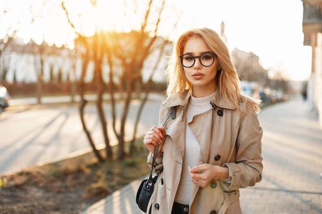 Hermosa mujer joven en gafas hipster con abrigo de moda y bolso caminando por la calle al atardecer