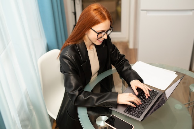 Hermosa mujer joven con gafas con el pelo rojo en la mesa con un portátil en la oficina