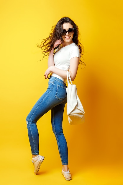 Hermosa mujer joven en gafas de sol, camisa blanca, jeans azul posando con bolsa