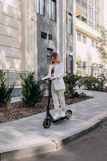 Hermosa mujer joven con gafas de sol y traje blanco está montando en su scooter eléctrico cerca de un edificio moderno en la ciudad y mirando hacia otro lado