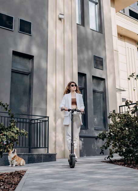 Hermosa mujer joven con gafas de sol y traje blanco está montando en su scooter eléctrico con perro Corgi cerca de un edificio moderno en la ciudad y mirando hacia otro lado