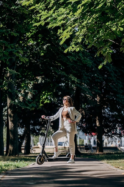 Hermosa mujer joven con gafas de sol y traje blanco está de pie con su scooter eléctrico en el parque de la ciudad