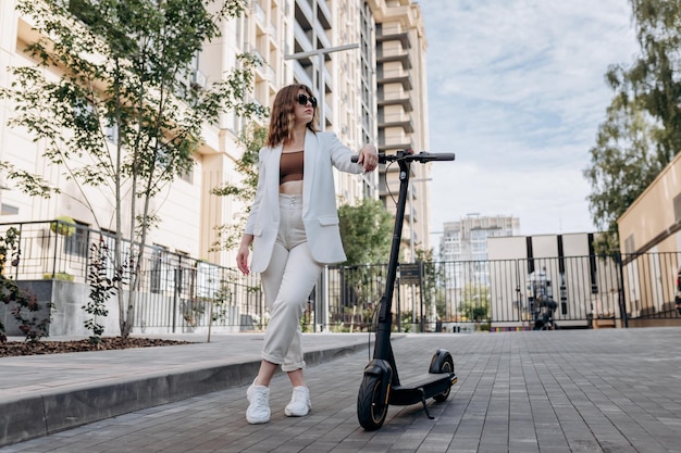 Hermosa mujer joven con gafas de sol y traje blanco parada con su scooter eléctrico cerca de un edificio moderno en la ciudad y mirando hacia otro lado