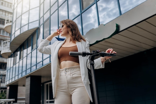 Hermosa mujer joven con gafas de sol y traje blanco de pie con su scooter eléctrico cerca de un edificio moderno y mirando hacia otro lado