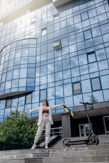 Foto hermosa mujer joven con gafas de sol y traje blanco de pie con su scooter eléctrico cerca de un edificio moderno y mirando hacia otro lado