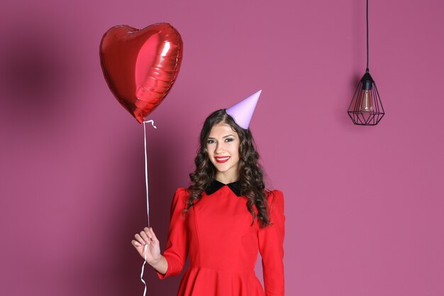 Hermosa mujer joven en gorra de fiesta con globo de aire en forma de corazón en la superficie de color