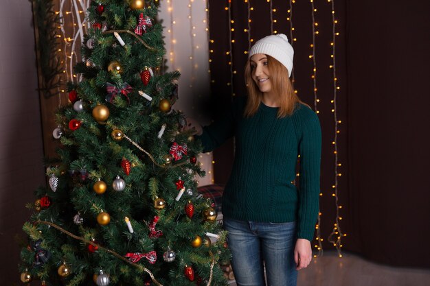 Hermosa mujer joven con una gorra y un suéter sonriendo y de pie cerca del árbol de Navidad