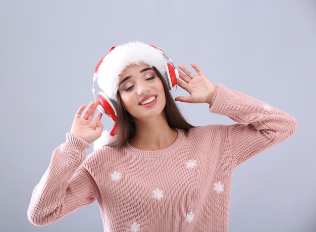 Hermosa mujer joven con gorro de Papá Noel escuchando música navideña en gris