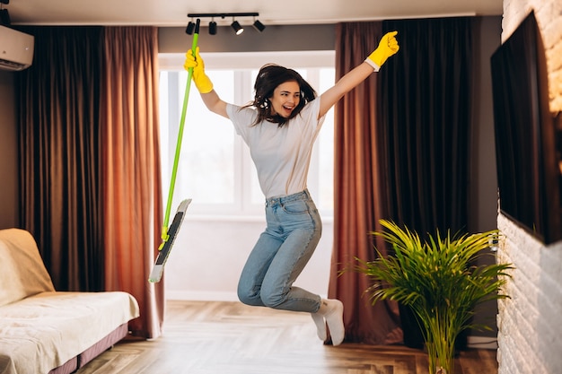 Foto hermosa mujer joven en guantes amarillos bailando con un trapeador mientras limpia la sala de estar.
