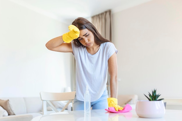 Hermosa mujer joven hace la limpieza de la casa Chica frota el polvo Mujer sonriente con guantes amarillos protectores de goma limpiando con un trapo y una botella de spray detergente Concepto de limpieza del hogar