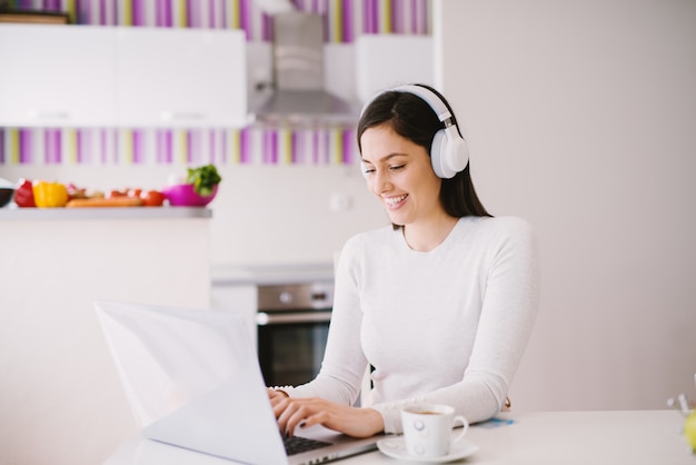 Hermosa mujer joven hermosa está utilizando su computadora portátil mientras escucha música en un auricular y tomando café en la habitación luminosa.