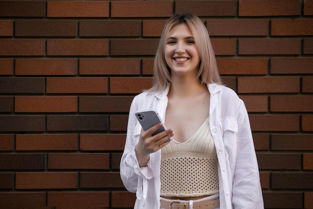 Hermosa mujer joven hermosa con mensajes de cabello rubio en el teléfono inteligente en el fondo de la calle de la ciudad chica guapa que tiene una conversación de teléfono inteligente en la calle de la ciudad