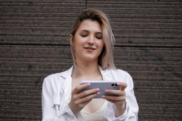 Hermosa mujer joven hermosa con mensajes de cabello rubio en el teléfono inteligente en el fondo de la calle de la ciudad chica guapa que tiene una conversación de teléfono inteligente en la calle de la ciudad