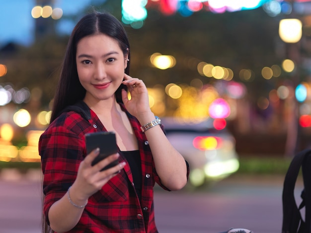 Hermosa mujer joven hermosa con smartphone en la calle de la ciudad, luz de noche borrosa en segundo plano.