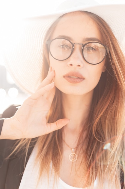 Hermosa mujer joven con hermosos labios en gafas de moda con sombrero elegante elegante blanco de paja en chaqueta de moda negra posando en la luz del sol brillante. Encantadora chica guapa al atardecer. Retrato de moda.