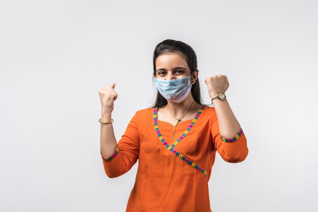 Hermosa mujer joven india sonriente en mascarilla protectora, feliz mujer milenaria en cubierta facial médica de pensamiento COVID-19, concepto de salud, aislado sobre pared blanca