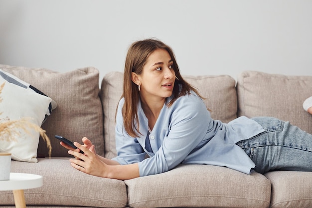 Hermosa mujer joven en jeans y camisa azul acostada en el sofá con el teléfono en la mano