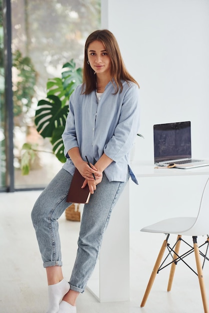 Hermosa mujer joven en jeans y camisa azul de pie junto a la mesa con el portátil en el interior