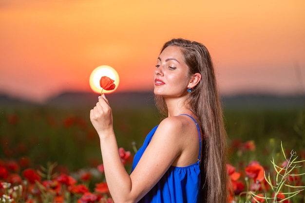Hermosa mujer joven jugando con la bola del sol mientras está de pie en el campo de amapolas en la cálida luz del atardecer