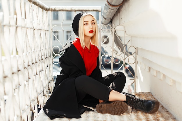 Hermosa mujer joven con labios rojos en ropa de otoño negro de moda sentado cerca de la barandilla de metal