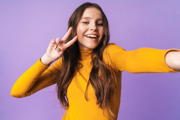 Hermosa mujer joven con largo cabello castaño gesticulando y tomando selfie foto celular aislado