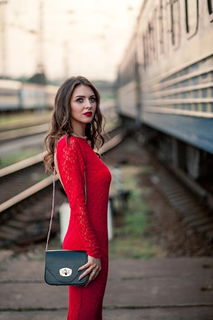 Hermosa mujer joven con un largo vestido rojo se encuentra en la plataforma de la estación cerca del ferrocarril