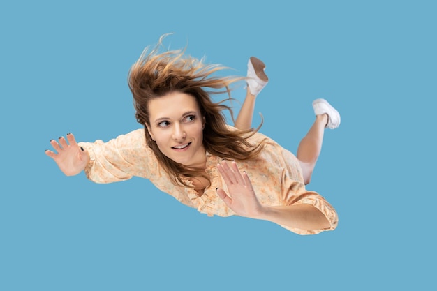 Hermosa mujer joven levitando en el aire, cayendo y su cabello desordenado volando por el viento, modelo volando flotando con una expresión pacífica de ensueño. Tiro interior aislado sobre fondo azul.