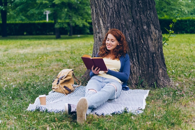 Hermosa mujer joven leyendo un libro sentado en una manta bajo un árbol en el parque sonriendo