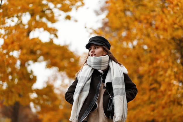 Hermosa mujer joven linda con estilo en una chaqueta marrón de moda en un elegante sombrero negro con una bufanda cálida vintage camina en el parque de otoño. Modelo de moda atractiva chica europea disfruta de un paseo al aire libre.