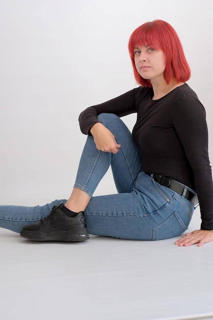 Hermosa mujer joven linda con peinado naranja en jeans sentada posando en el estudio sobre fondo blanco