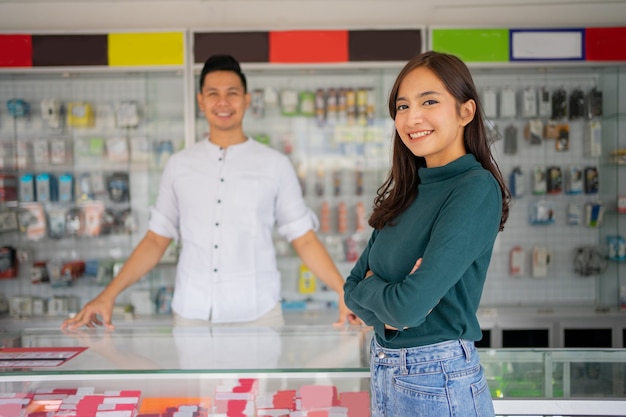 Hermosa mujer joven con las manos cruzadas de pie contra una tienda de teléfonos inteligentes