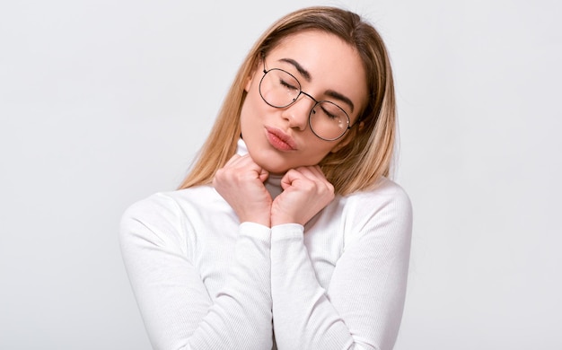 Hermosa mujer joven mantiene las manos en el pecho con los ojos cerrados usando blusa blanca y anteojos redondos aislados sobre fondo blanco Personas emociones sentimiento y concepto de lenguaje corporal