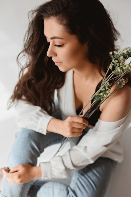 Hermosa mujer joven con maquillaje nude con jeans y blusa desabrochada posando con una ramita de f ...