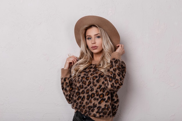 Hermosa mujer joven maravillosa con hermosos ojos con labios sexy en un elegante sombrero beige con pantalones de cuero en un suéter de leopardo vintage posando