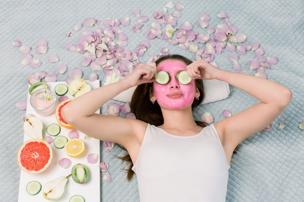 Foto hermosa mujer joven con máscara facial en la cara con rodajas de pepino fresco.