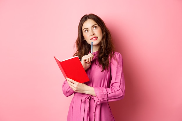 Hermosa mujer joven mira pensativa, escribiendo en un cuaderno, sosteniendo un planificador o un diario, planifica su horario, de pie contra la pared rosa. Copia espacio