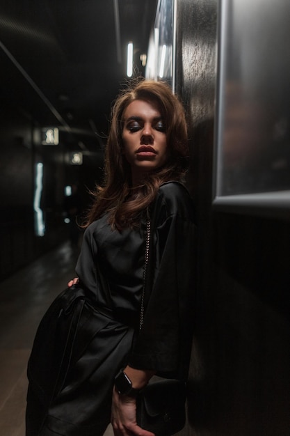 Hermosa mujer joven de moda en un elegante vestido negro de moda se encuentra cerca de la pared en el interior oscuro