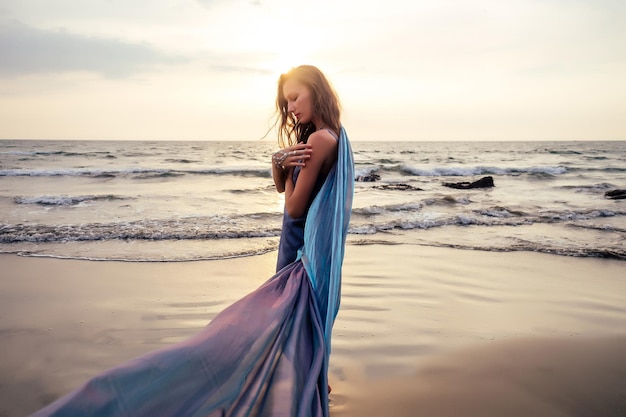 Hermosa mujer joven morena de estilo en azul camaleón vestido largo tren de pie sobre una roca cerca del mar, arena tropical sobre una roca.hermosa sirena solitaria melancolía esperando libertad y autónomo