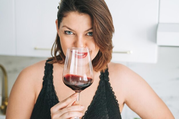 Foto hermosa mujer joven morena sensual feliz cuerpo de talla grande positivo con una copa de vino tinto en casa