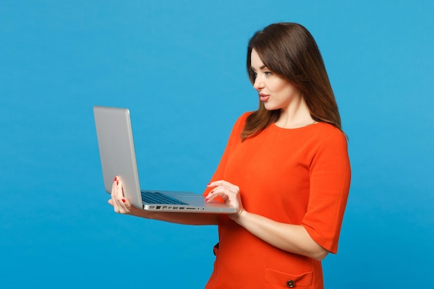 Hermosa mujer joven morena con vestido rojo anaranjado sostenga en las manos usando una computadora portátil aislada en el fondo de la pared azul, retrato de estudio. Concepto de moda de estilo de vida de la gente. Simulacros de espacio de copia.