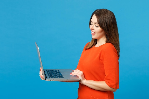 Hermosa mujer joven morena con vestido rojo anaranjado sostenga en las manos usando una computadora portátil aislada en el fondo de la pared azul, retrato de estudio. Concepto de moda de estilo de vida de la gente. Simulacros de espacio de copia.