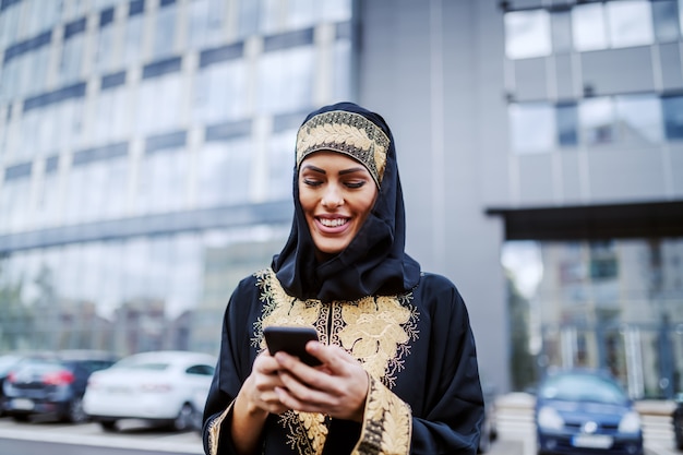 Hermosa mujer joven musulmana positiva sonriente de pie frente al edificio corporativo y usando teléfonos inteligentes para enviar un correo electrónico. Generación millennial.