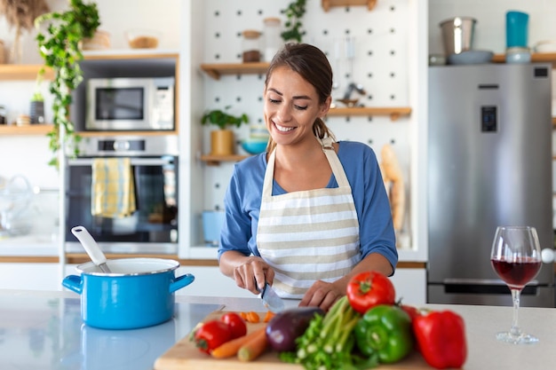 Hermosa mujer joven parada en la cocina moderna cortar verduras preparar ensalada de verduras frescas para la cena o el almuerzo mujer joven cocinando en casa hacer el desayuno seguir una dieta saludable concepto vegetariano