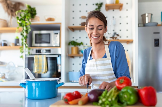 Hermosa mujer joven parada en la cocina moderna cortar verduras preparar ensalada de verduras frescas para la cena o el almuerzo mujer joven cocinando en casa hacer el desayuno seguir una dieta saludable concepto vegetariano