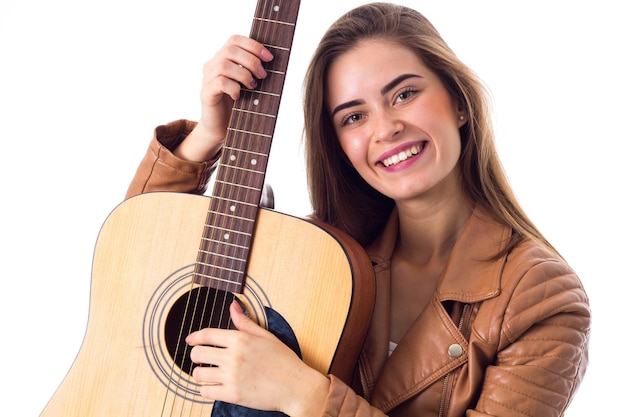 Hermosa mujer joven con el pelo largo en chaqueta marrón sosteniendo una guitarra sobre fondo blanco en el estudio